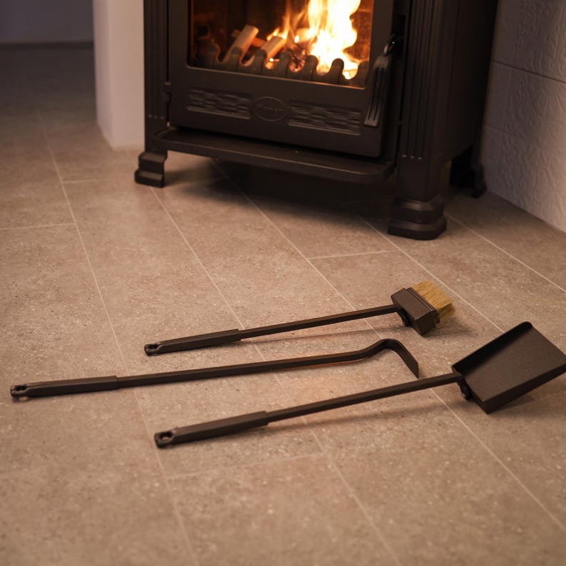May include: A set of three fireplace tools: a poker, a brush, and a shovel. The tools are made of black metal and have wooden handles. The tools are lying on a light brown tiled floor in front of a black wood-burning stove.
