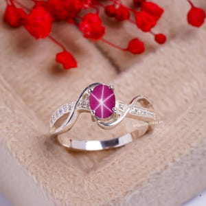 May include: A silver ring with a pink star sapphire gemstone and a row of small clear gemstones on each side. The ring is set in a twisted band.