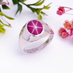 Op de afbeelding: Een zilveren ring met een roze ster-saffier edelsteen. De ring heeft een afgeronde band en de edelsteen is gezet in een bezel setting. De ring is gegraveerd met de letters "SF".
