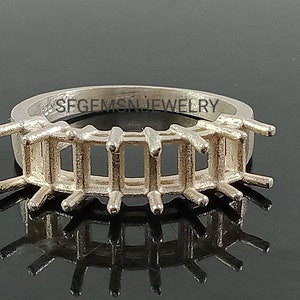 May include: Silver ring setting with a row of five emerald-cut gemstone settings. The setting is designed to hold five emerald-cut gemstones in a row. The ring is made of silver and has a polished finish. The setting is designed to hold gemstones that are approximately 5mm x 7mm in size. The ring is marked "SFGEMSNJEWELRY".