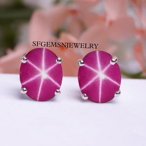 May include: A pair of silver stud earrings featuring pink star ruby gemstones. Each gemstone has a six-pointed star design in the centre.