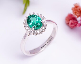 May include: A silver ring with a large, oval emerald-green gemstone, encircled by smaller, clear stones. The ring's band is polished, with the text "SFGEMSNJWELRY" engraved on the side. A classic piece of jewellery.