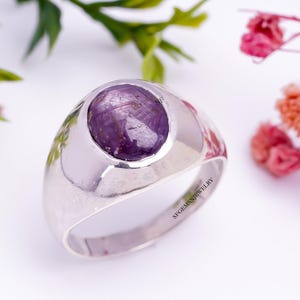 Può includere: Un anello d'argento con una pietra preziosa ovale di colore viola incastonata in una montatura rialzata. L'anello è inciso con il testo "SFGEMS JEWELRY".
