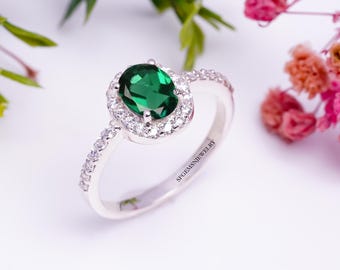 May include: A silver ring featuring a large oval green gemstone encircled by a halo of smaller white gemstones. The ring band is adorned with smaller white gemstones.