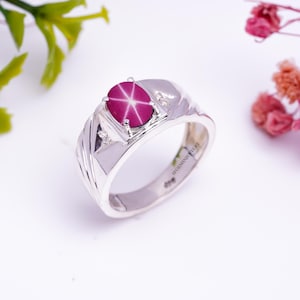 Puede incluir: Un anillo de plata con una piedra preciosa de rubí estrellado rosa y dos diamantes más pequeños. El anillo está grabado con "SG GEMSNIF JEWELRY".