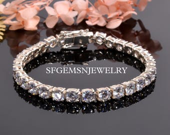 May include: A silver tennis bracelet with a row of clear round cubic zirconia stones. The bracelet is on a black reflective surface.