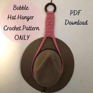 May include: A pink crocheted hat hanger with a wooden ring at the top. The hanger is hanging on a wall with a brown hat hanging from it. The text "PDF Download" is visible in the upper right corner of the image.