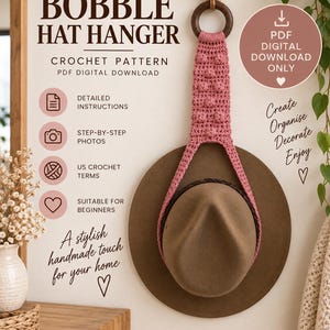 May include: A crochet pattern for a bobble hat hanger, a PDF digital download. The design features a pink crocheted strap with bobble texture attached to a dark wooden ring, holding a brown felt hat. Text includes Bobble Hat Hanger.