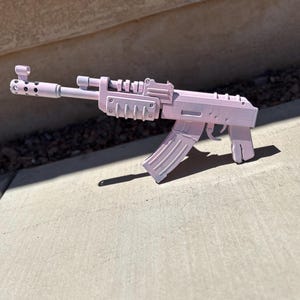May include: A pink AK-47 style toy gun. The toy gun is made of plastic and has a magazine, trigger, and sight. The gun is painted a solid light pink color and is resting on a concrete surface.