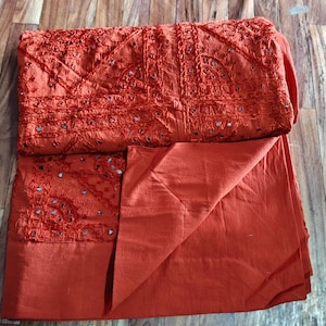 May include: A rust-coloured cotton throw blanket with intricate embroidery and mirror work. The blanket is folded in half and has a textured surface.