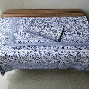 May include: A white tablecloth with a blue floral pattern and a blue border. The tablecloth is folded in half and placed on a wooden table.