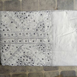 May include: White cotton fabric with silver embroidery and sequins. The fabric has a geometric pattern with a border of plain white fabric.