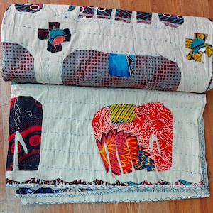 Könnte beinhalten: Eine weiße Baumwolldecke mit bunten Stoffapplikationen. Die Decke zeigt eine Vielzahl von Mustern und Farben, darunter Rot, Gelb, Blau und Grün. Die Decke ist in der Hälfte gefaltet und die Kanten sind mit einem Zickzack-Stich vernäht.