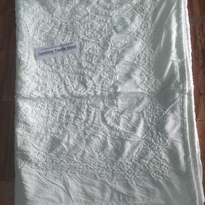 May include: A white cotton fabric with an embossed floral pattern. The fabric is folded and has a tag that says "Sunshine Textile Store".