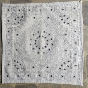 May include: A white square cushion with a decorative embroidered design featuring a central diamond shape and a circle of small black mirrors. The cushion has a white border with a repeating pattern.