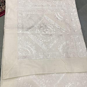 Pode incluir: Uma toalha de mesa branca bordada com um delicado padrão floral. A toalha de mesa é quadrada e tem um design simples.