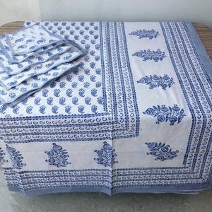May include: A white tablecloth with a blue paisley pattern and a blue border. The tablecloth is folded and has a stack of matching napkins on top.