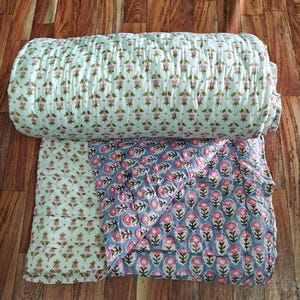 May include: A quilted blanket featuring a white base with a repeating pattern of small pink flowers and green leaves, while the underside displays a blue background with a larger repeating pattern of pink floral motifs and green stems.