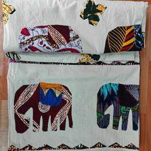 May include: A white fabric with a patchwork design featuring colourful elephants. The elephants are made from different patterned fabrics, including floral, geometric, and abstract designs. The fabric has a brown border with a zig-zag pattern.