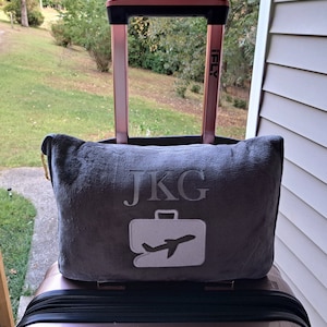 Puede incluir: Una almohada de viaje gris con las iniciales "JKG" y un icono de avi&oacute;n blanco bordado en ella. La almohada est&aacute; encima de una maleta de color rosa dorado con un asa negra.