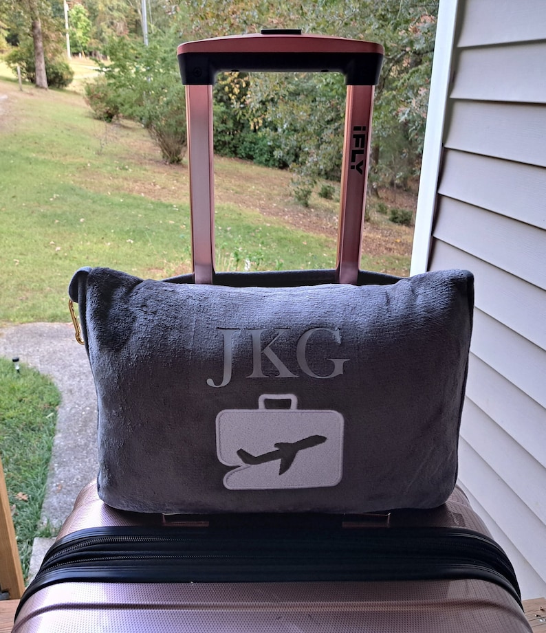 Puede incluir: Una almohada de viaje gris con las iniciales "JKG" y un icono de avi&oacute;n blanco bordado en ella. La almohada est&aacute; encima de una maleta de color rosa dorado con un asa negra.
