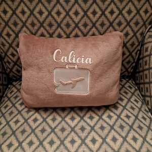 Puede incluir: Una almohada de felpa marr&oacute;n con el nombre "Calicia" bordado. La almohada tiene un dise&ntilde;o de avi&oacute;n y maleta blancos.