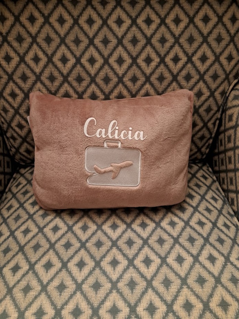 Puede incluir: Una almohada de felpa marr&oacute;n con el nombre "Calicia" bordado. La almohada tiene un dise&ntilde;o de avi&oacute;n y maleta blancos.