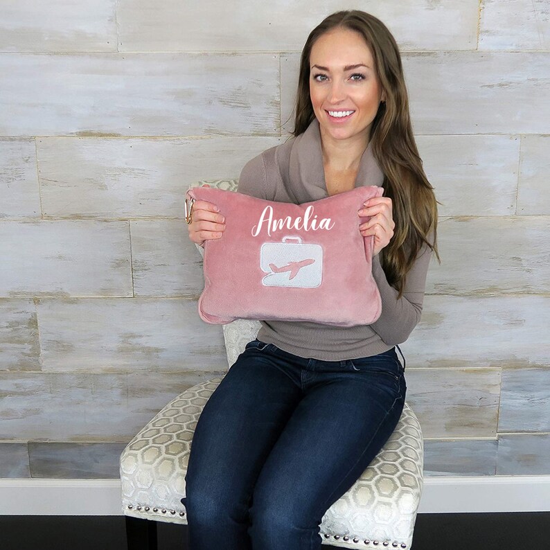Puede incluir: Una almohada de viaje rosa con el nombre "Amelia" bordado. La almohada tambi&eacute;n presenta un avi&oacute;n y una maleta blancos.
