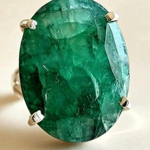 May include: A ring featuring a large, oval-cut, deep green gemstone with visible internal inclusions and facets. The stone is secured in a four-prong setting made of a polished, silver-toned metal band that extends from the base of the setting.