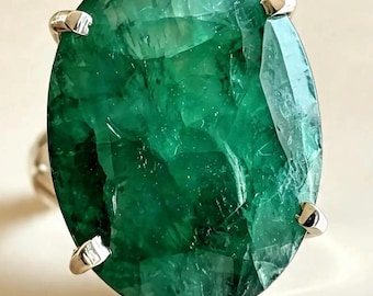 May include: A ring featuring a large, oval-cut, deep green gemstone with visible internal inclusions and facets. The stone is secured in a four-prong setting made of a polished, silver-toned metal band that extends from the base of the setting.