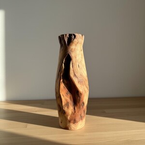 May include: A handcrafted wooden vase with a natural, organic shape. The vase is made from light brown wood with darker brown knots and grain patterns. The top of the vase has a textured, uneven edge. The vase is displayed on a light-colored wooden surface.