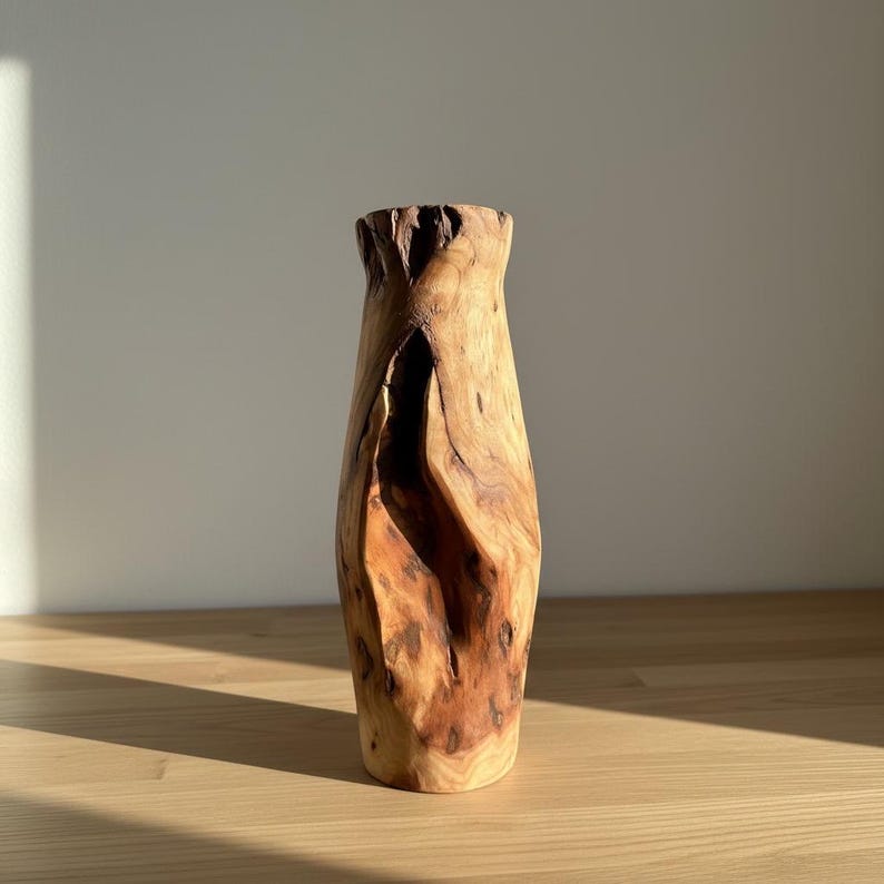 May include: A handcrafted wooden vase with a natural, organic shape. The vase is made from light brown wood with darker brown knots and grain patterns. The top of the vase has a textured, uneven edge. The vase is displayed on a light-colored wooden surface.