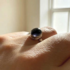 May include: A silver ring featuring a smooth, oval-shaped dark purple gemstone set in a simple metal bezel. The stone has a polished, reflective surface that catches the light, and the band is a plain, solid silver metal construction.