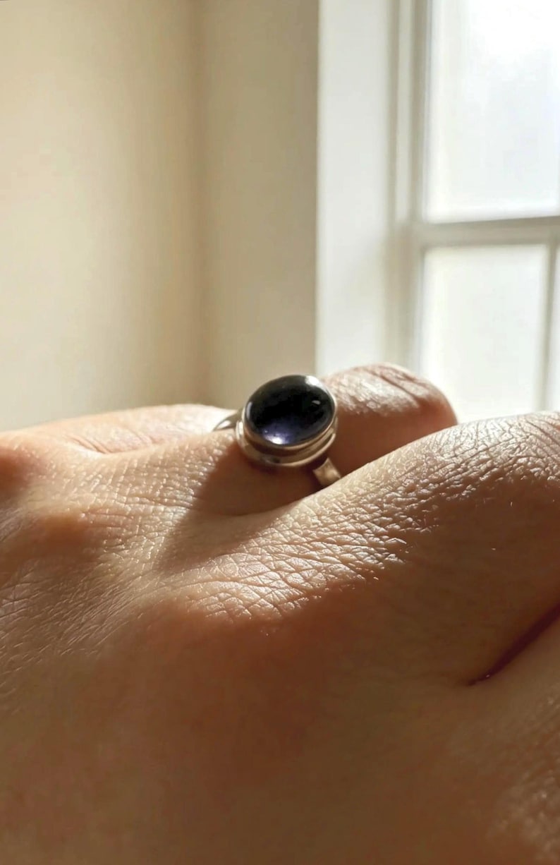 May include: A silver ring featuring a smooth, oval-shaped dark purple gemstone set in a simple metal bezel. The stone has a polished, reflective surface that catches the light, and the band is a plain, solid silver metal construction.