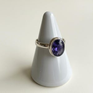 May include: A silver ring featuring a large, oval-shaped, deep purple faceted gemstone set in a smooth metal bezel. The ring is displayed on a white conical stand, highlighting the polished silver band and the dark violet stone's clarity.