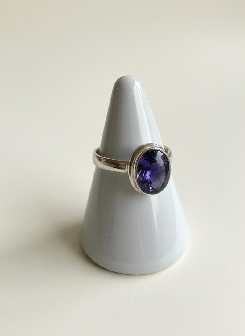 May include: A silver ring featuring a large, oval-shaped, deep purple faceted gemstone set in a smooth metal bezel. The ring is displayed on a white conical stand, highlighting the polished silver band and the dark violet stone's clarity.