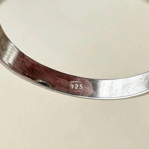 May include: A silver-coloured metal ring band featuring a smooth, polished finish. The interior surface of the band is clearly stamped with the hallmark 925, indicating the metal composition. The ring has a simple, curved, and continuous circular shape.