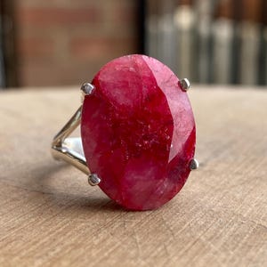 May include: A silver ring featuring a large, oval-shaped ruby gemstone. The ruby is a vibrant red hue. The ring is presented on a wooden surface. The gemstone is approximately 2.5 cm in length.