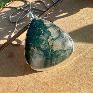 May include: A silver pendant necklace with a triangular, translucent stone containing green, moss-like patterns. The pendant is set in a silver bezel and suspended from a fine silver chain. The necklace is displayed on a light brown surface.