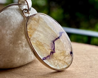 May include: Oval pendant with a silver-colored metal frame. The pendant features a light-colored stone with gold and purple veining, suspended from a silver chain. The pendant is displayed on a white stone.