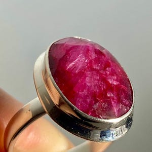 May include: A ring featuring a large, oval-shaped, deep pinkish-red faceted gemstone set in a smooth, polished silver-toned metal bezel. The stone displays a textured, crystalline surface with natural inclusions and a vibrant, saturated colour.