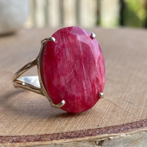 May include: A silver ring with a large, oval-cut ruby gemstone. The ruby is a bright red colour with internal patterns. The ring has a simple split-shank band, and the gemstone is secured by four prongs. The ring is set on a wooden surface.