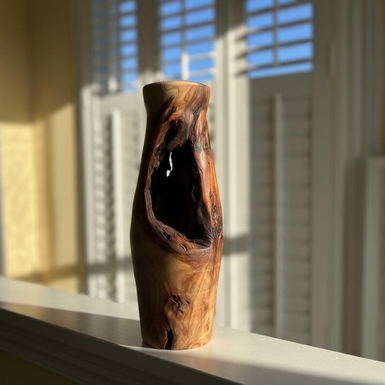 May include: A tall, handcrafted wooden vase with a natural, organic design. The vase features a large, irregular opening in the center, revealing a dark interior. The wood grain is visible, showcasing shades of brown and tan. The vase is displayed on a white surface.