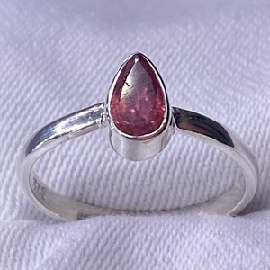 May include: A silver ring with a pear-shaped, reddish-pink gemstone. The ring has a simple, elegant design with a smooth, polished band. The gemstone is set in a bezel setting, enhancing its colour and shape. The ring is displayed on a white surface.