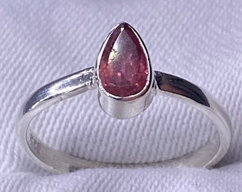 May include: A silver ring with a pear-shaped, reddish-pink gemstone. The ring has a simple, elegant design with a smooth, polished band. The gemstone is set in a bezel setting, enhancing its colour and shape. The ring is displayed on a white surface.