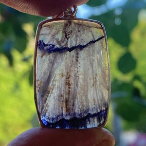 May include: A rectangular pendant with a silver-coloured frame. The pendant features a translucent stone with a mix of white, blue, and brown hues. The stone's interior has a textured, layered appearance, with dark blue streaks and patterns.