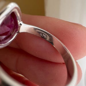 以下が含まれることがあります： 磨かれた金属バンドにセットされた、大きなファセットカットの濃いピンク色の宝石が付いたシルバーリング。バンドの内側には925の刻印がはっきりとあり、このジュエリーがスターリングシルバー製であることを示しています。