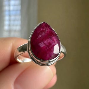 以下が含まれることがあります： 滑らかな金属製のベゼルにセットされた、大きな洋梨型のファセットカットが施された深いマゼンタ色のルビーの指輪。リング部分は磨かれたシルバーで、石の表面は自然な質感で、内部のインクルージョンやカット面がはっきりと見えています。