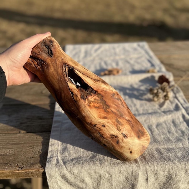 May include: A handcrafted wooden vase with a natural, organic shape. The vase is made from a light brown wood with darker brown and black grain patterns. It has a large opening and a hollowed-out interior, perfect for displaying flowers or as a decorative piece.