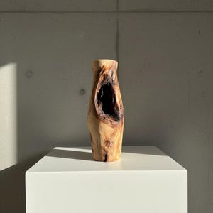 May include: A handcrafted wooden vase with a natural, organic shape. The vase is made from light brown wood and features a large, dark opening in the center. It stands on a white pedestal against a gray concrete wall.
